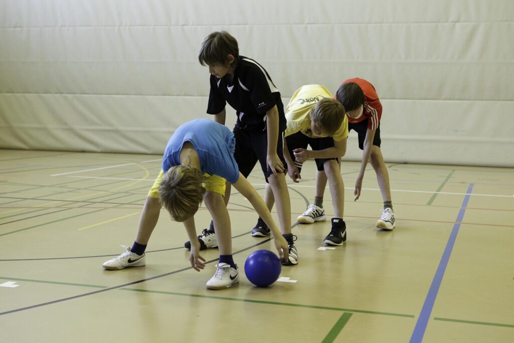 Vier Jungen stehen in einer Reihe in einer Turnhalle und beugen sich nach vorne, während der erste Junge einen blauen Ball durch seine Beine rollt.