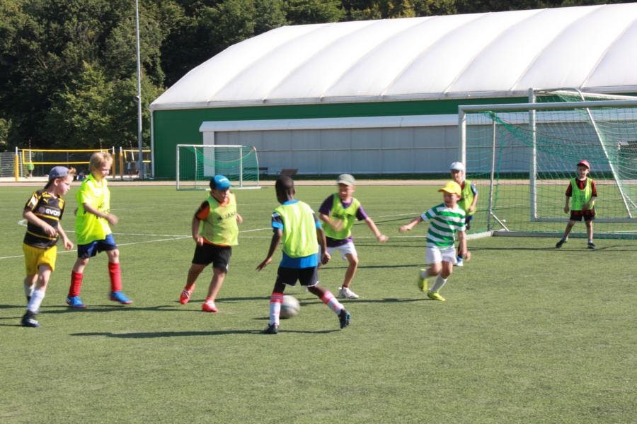 Mehrere Kinder spielen Fußball auf einem Kunstrasenplatz mit einer großen, weißen Sporthalle im Hintergrund.
