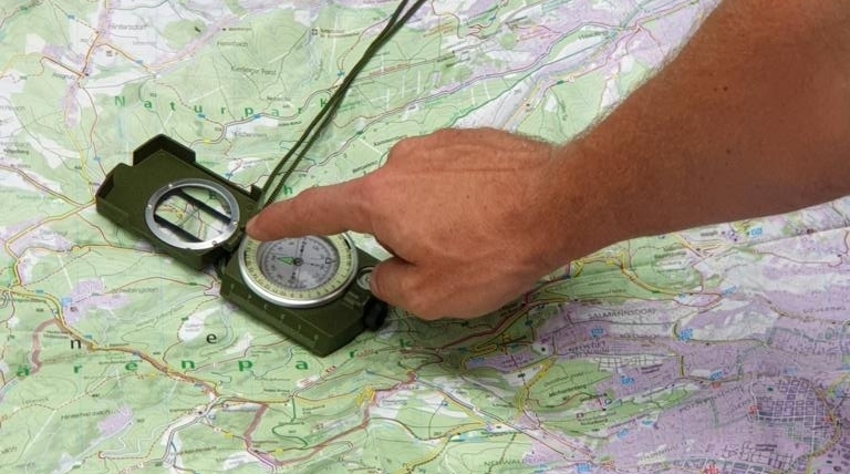 Eine Hand zeigt auf einen Marschkompass, der auf einer topografischen Karte liegt.