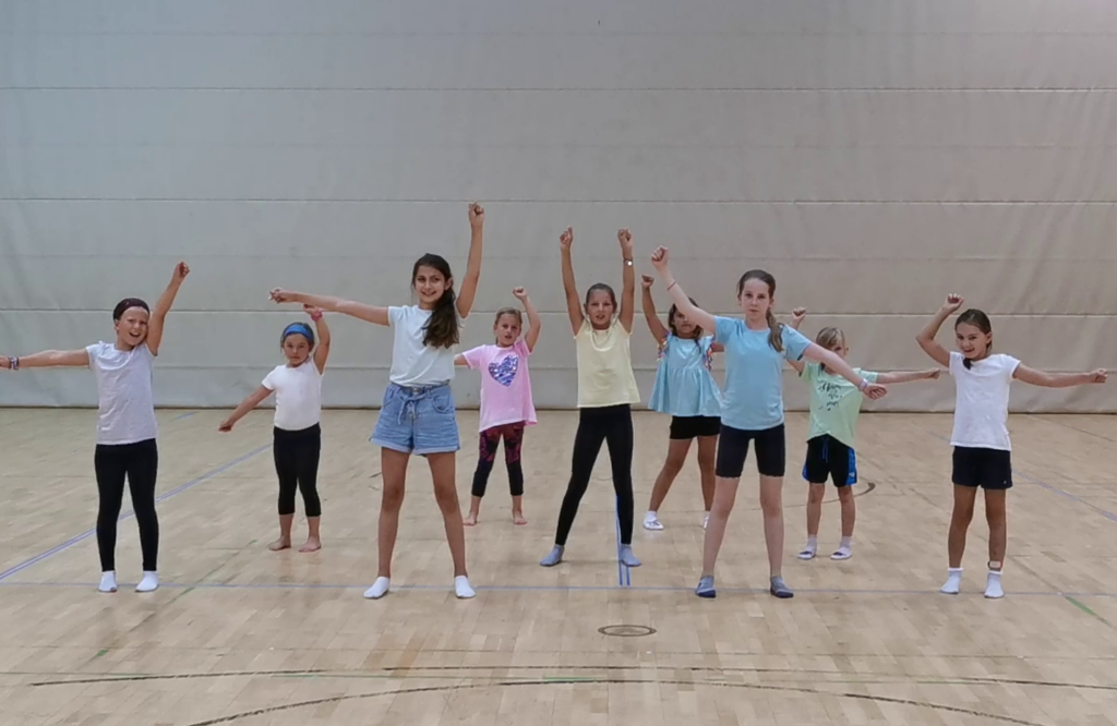 Eine Gruppe von Mädchen steht in einer Turnhalle in einer Linie, hebt eine Faust in die Luft und lächelt – eine tänzerische Pose.
